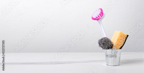 Tiny metal bucket and sponge, cleaning brush, scrubber in it. Cleaning tools on the white table against white wall.Empty space for text