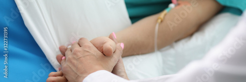 Close-up view of doctor in medical gown holding patients hands. Sick person laying with drop bottle and catheter. Healthcare and medicine treatment concept