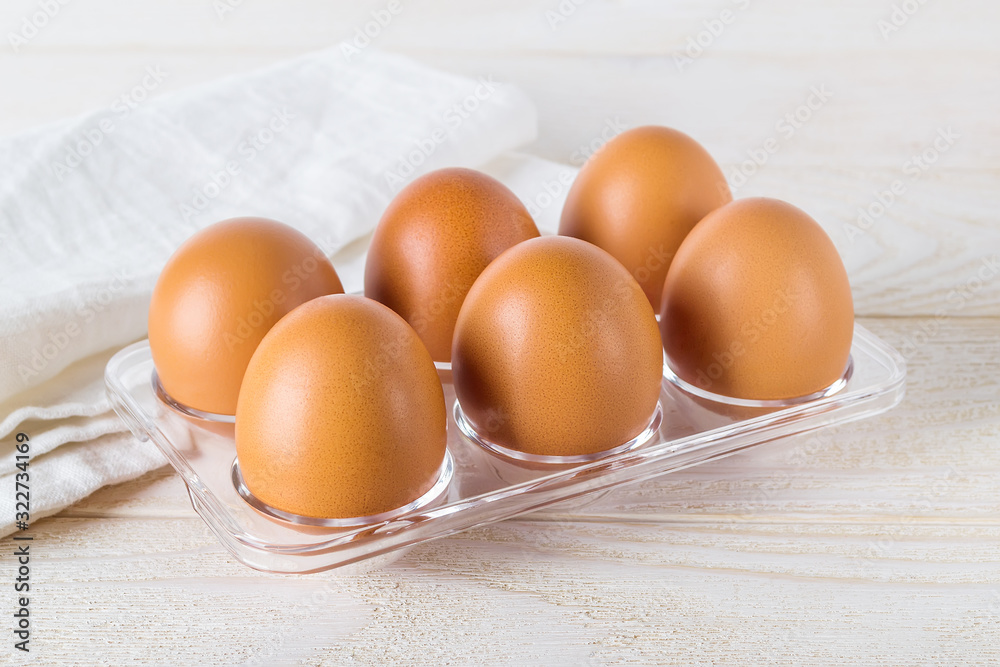 Six raw chicken eggs in a transparent plastic tray on a white wooden table. Half a dozen brown eggs prepared for cooking. Cooking for easter. Six raw chicken eggs in a transparent plastic tray on a white wooden table. Half a dozen brown eggs prepared for cooking. Cooking for easter.