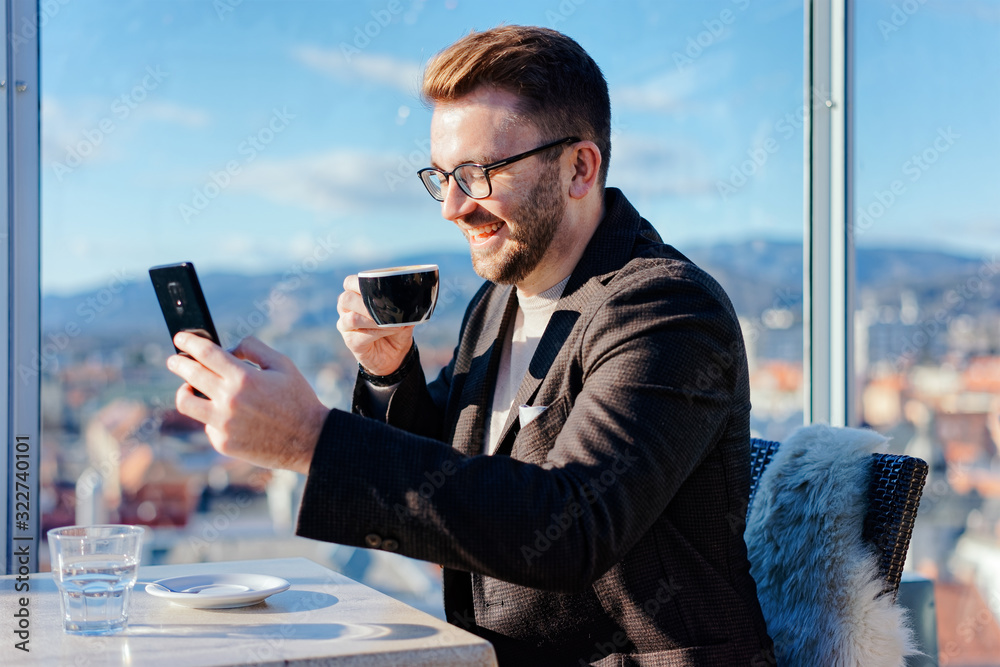 One caucasian man wearing glasses holds smartphone while drinks coffee from cup in cafe
