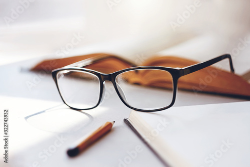 On a white table lies an open book, elegant black rimmed glasses, a notebook for notes and a pencil, illuminated by the light of the midday sun.