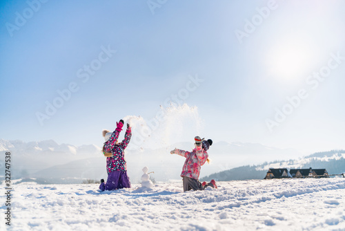 Cheerful little girls in outwear having fun and throwing snow in sunlight standing outdoors