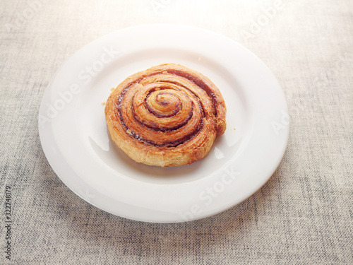 One tasty cinnamon roll on a white plate. Bakery product on a simple table cloth.