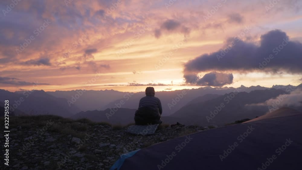 Camper in jacket and sleeping bag watching the sunset in mountain landscape Camper in jacket and sleeping bag watching the sunset in mountain landscape