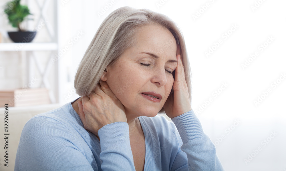 Woman suffering from stress or a headache grimacing in pain as she holds the back of her neck with her other hand to her temple, with copyspace. Concept photo with indicating location of pain. Woman suffering from stress or a headache grimacing in pain as she holds the back of her neck with her other hand to her temple, with copyspace. Concept photo with indicating location of pain.