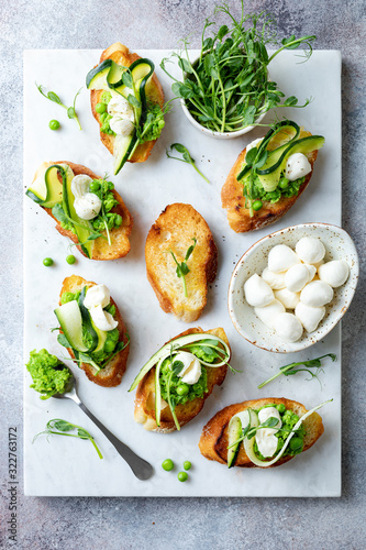 Appetizer crostini with mashed green pea, mozzarella, pea sprouts and zucchini ribbons on white marble board. Delicious healthy snack, spring appetizers