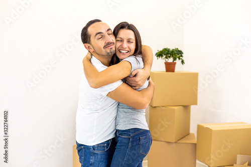 Smiling couple standing together, embracing in their new apartments with piles of boxes on the background. © Andrii