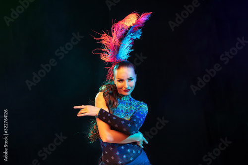 Moving. Beautiful young woman in carnival, stylish masquerade costume with feathers on black background in neon light. Copyspace for ad. Holidays celebration, dancing, fashion. Festive time, party.