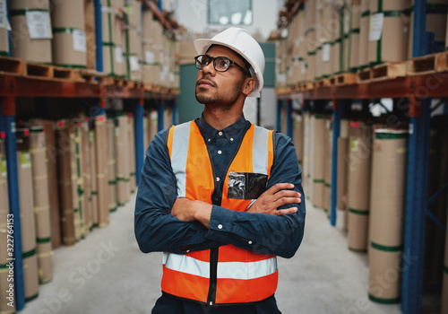 Confident warehouse supervisor in aisle standing with folded hands looking high at shelves with material while wearing orange vest and white hardhat
