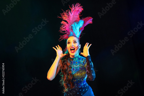 Surprised. Beautiful young woman in carnival, stylish masquerade costume with feathers on black background in neon light. Copyspace for ad. Holidays celebration, dancing, fashion. Festive time, party.