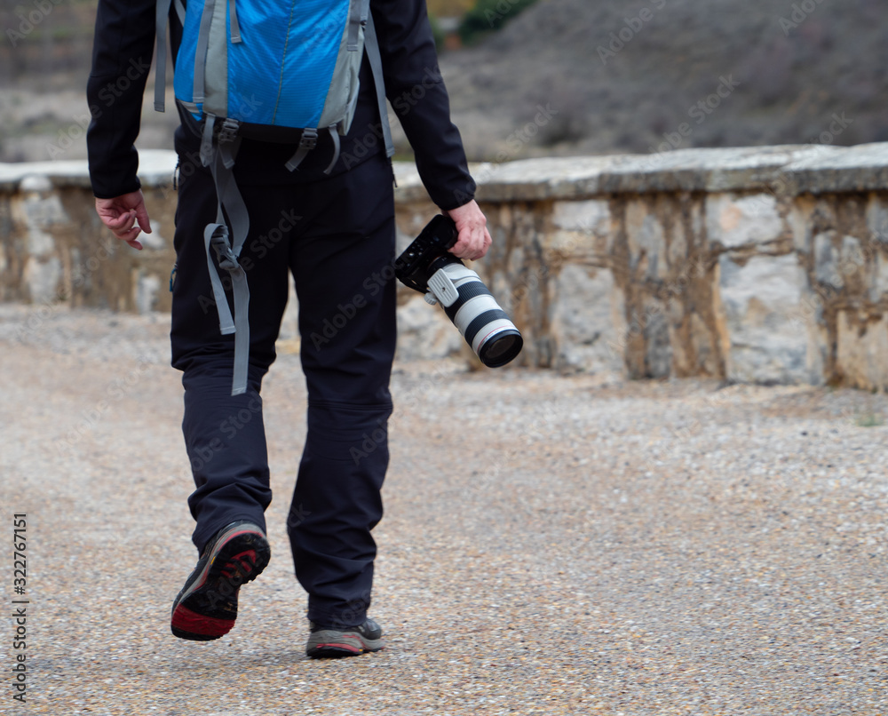 Man holding a camera with a big zoom objective outdoors in the middle of a path surrounded by a stone wall