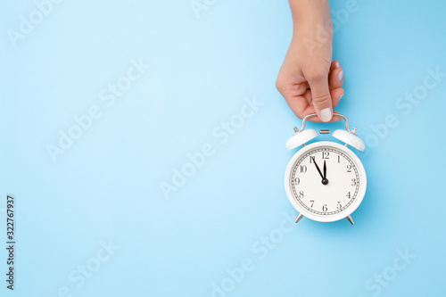 Woman hand holding white alarm clock on light blue table background. Pastel color. Time concept. Closeup. Empty place for text, quote or sayings. Top down view.