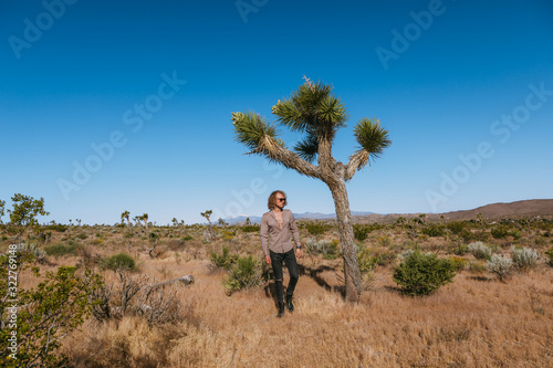 Joshua tree a curly haired blonde man is walking in the fierce heat and deep blue skies of the south western desert of North America, wearing dark sunglasses, a beige linen shirt