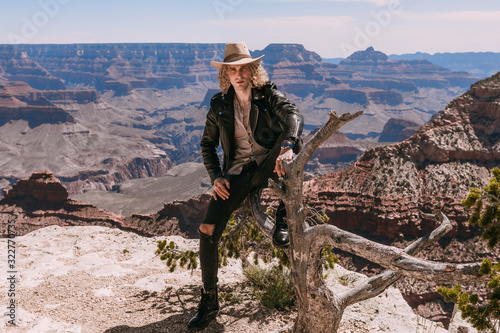 A Curly haired blonde man  wearing a black leather jacket  black jeans  black shoes  beige linen shirt and matching cowboy hat  perched on an old tree stump with background vistas of the Grand Canyon