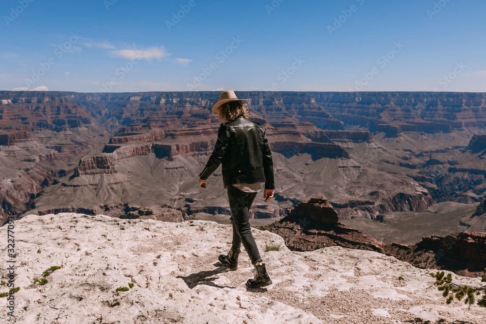 A Curly haired blonde man, wearing a black leather jacket ,black jeans, black shoes, beige linen shirt and matching cowboy hat, background vistas of the Grand Canyon