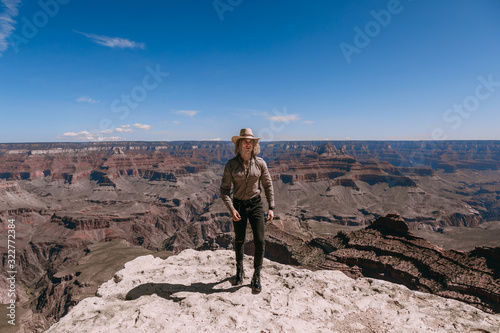 A Curly haired blonde man, wearing a beige T-Shirt ,black jeans, black shoes, beige linen shirt and matching cowboy hat, background vistas of the Grand Canyon © Georgi