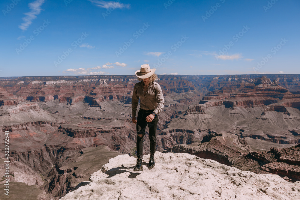 Fototapeta premium A Curly haired blonde man, wearing a beige T-Shirt ,black jeans, black shoes, beige linen shirt and matching cowboy hat, background vistas of the Grand Canyon