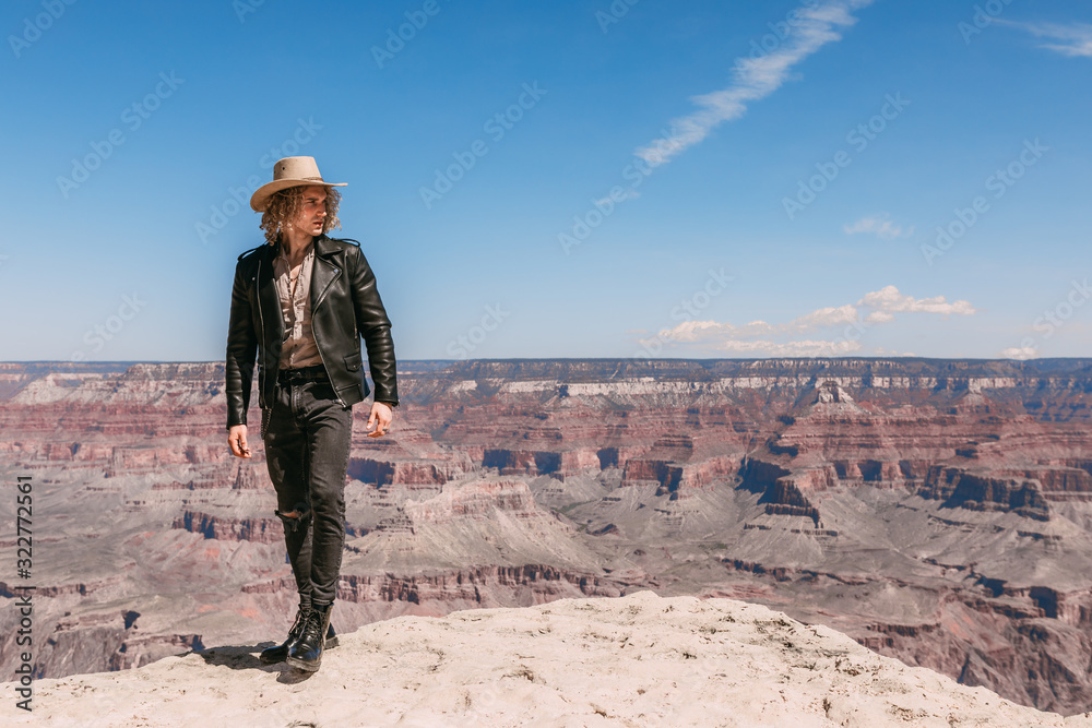 A Curly haired blonde man, wearing a black leather jacket ,black jeans, black shoes, beige linen shirt and matching cowboy hat, background vistas of the Grand Canyon A Curly haired blonde man, wearing a black leather jacket ,black jeans, black shoes, beige linen shirt and matching cowboy hat, background vistas of the Grand Canyon
