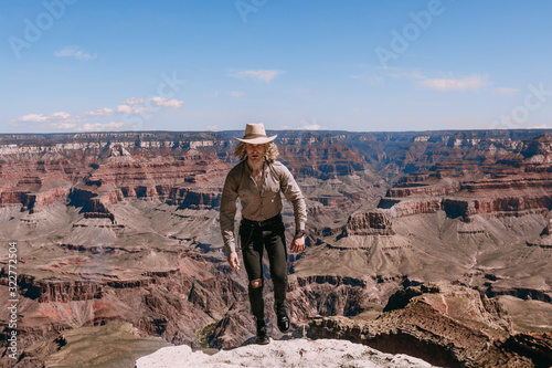A Curly haired blonde man, wearing a beige T-Shirt ,black jeans, black shoes, beige linen shirt and matching cowboy hat, background vistas of the Grand Canyon © Georgi