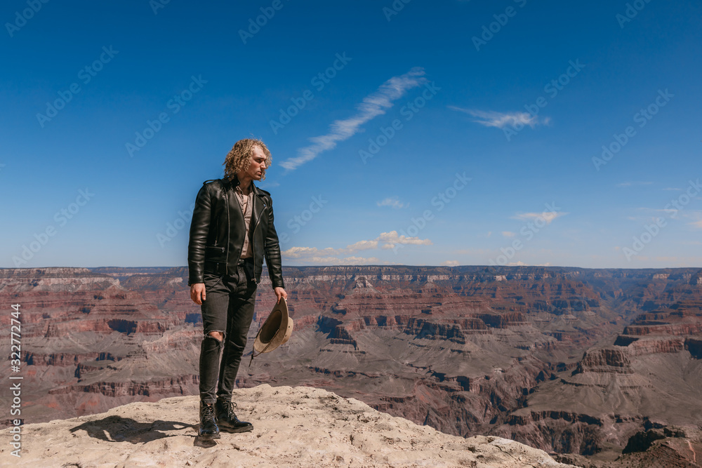 Fototapeta premium A Curly haired blonde man, wearing a black leather jacket ,black jeans, black shoes, beige linen shirt and matching cowboy hat, background vistas of the Grand Canyon