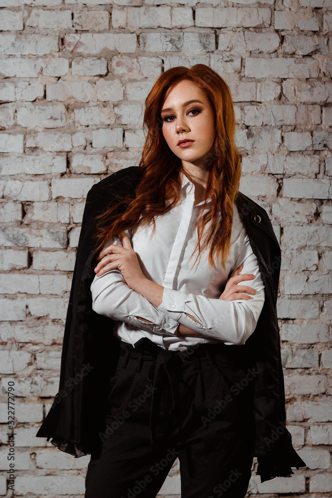 Young red-haired lady in a black suit consisted of trousers and jacket stands and looks directly. Self-confident business woman. White brick background. Young red-haired lady in a black suit consisted of trousers and jacket stands and looks directly. Self-confident business woman. White brick background.