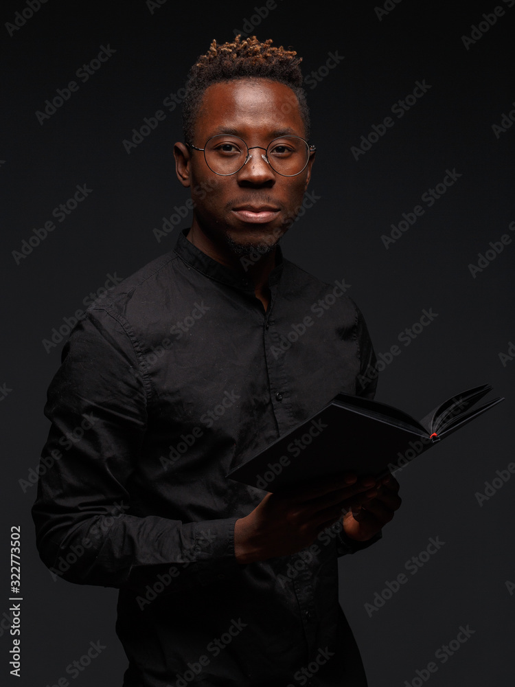 A young African man in black clothes on a dark background holds in the hands of a book. - obrazy, fototapety, plakaty 