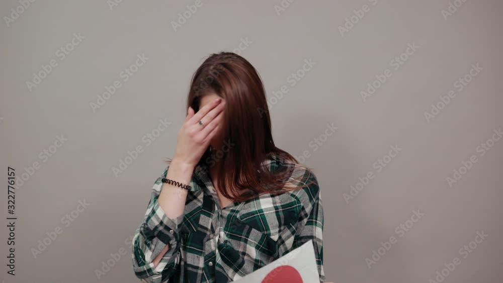 Young brunette girl blue green in checked shirt on grey background, an unhappy woman with glasses holds piece of paper with red heart in hand Young brunette girl blue green in checked shirt on grey background, an unhappy woman with glasses holds piece of paper with red heart in hand