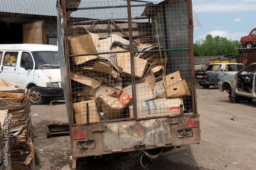 A trailer with cardboard boxes prepared for recycling stands in a landfill among old cars.