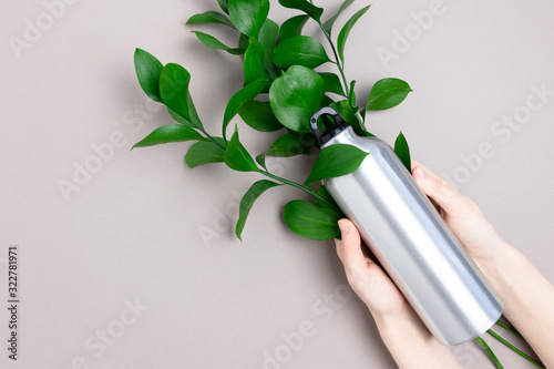 Hands hold metal bottle. Zero waste concept with bouquer of green leaves on gray background.