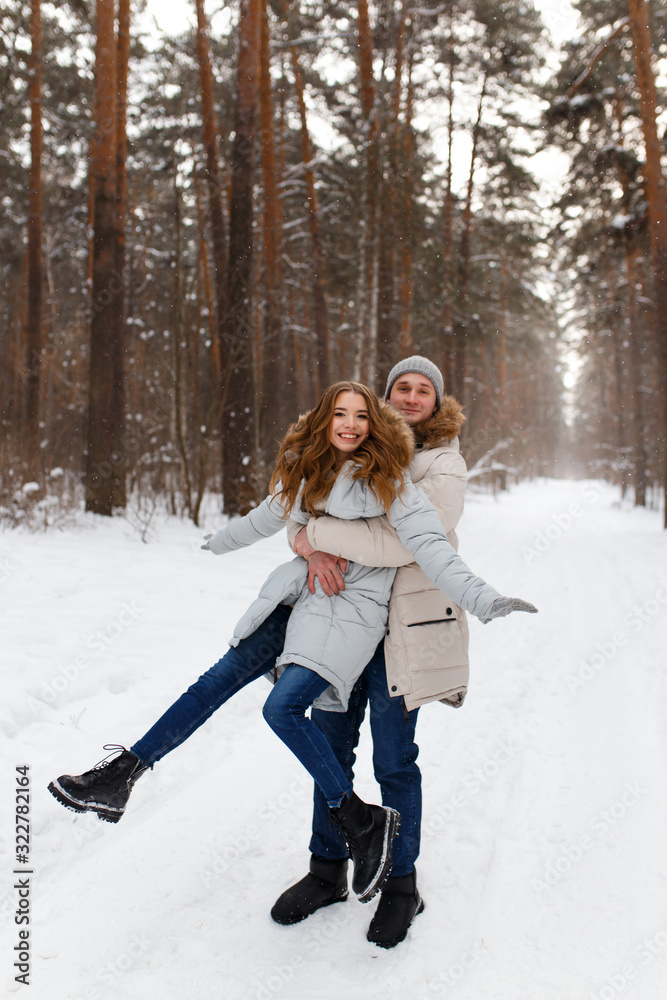 Fototapeta premium Happy lovers a guy and a girl who love each other hug, kiss, laugh, rage and walk in warm jackets in winter against the background of a snowy forest, a friendly family fun walk