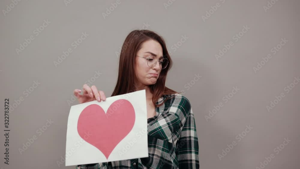 Young brunette girl blue green in checked shirt on grey background an offended woman holds piece of paper with a red heart, a negative attitude in her hand Young brunette girl blue green in checked shirt on grey background an offended woman holds piece of paper with a red heart, a negative attitude in her hand