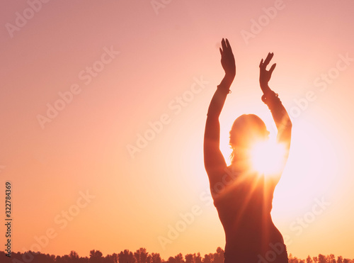 dark glowing silhouette of slim woman with hands up in the air illuminated with sunshine dancing traditional tribal belly dance in front of aurora sky at sunrise