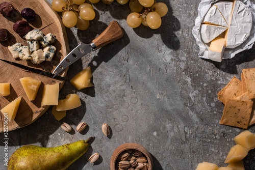 Top view of cheese, fruits, nuts, crackers and olives with knife and cutting board on grey background