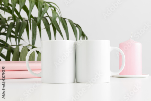 Two mugs mockup with books and a candle on a white table and a palm plant.