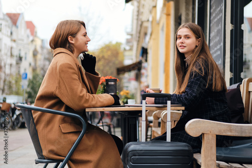Two beautiful stylish girls happily drinking coffee together while sitting in street cafe with suitcase