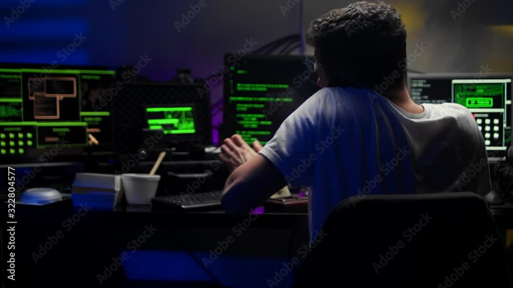 Close up of the Caucasian young handsome male virtual spy or hacker in a hood talking cheerfully on the phone while working at the computer in the dark room. Close up of the Caucasian young handsome male virtual spy or hacker in a hood talking cheerfully on the phone while working at the computer in the dark room.