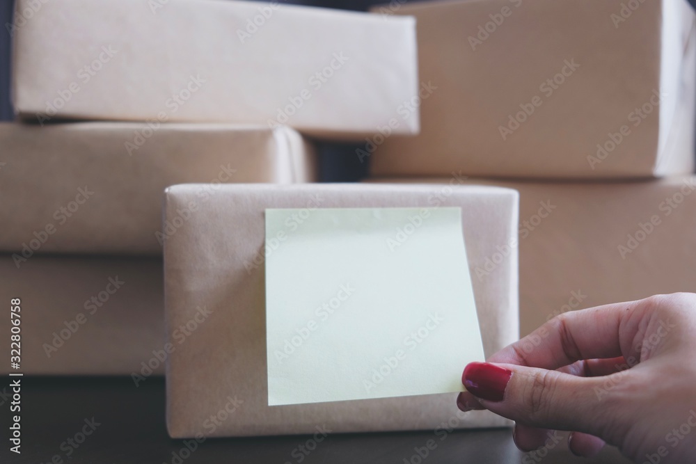 The small note attached on the product box wrapped by natural brown paper is captured or pulled by the hand of Asian woman. The stack of box are prepare to send or delivery product or gift to consumer The small note attached on the product box wrapped by natural brown paper is captured or pulled by the hand of Asian woman. The stack of box are prepare to send or delivery product or gift to consumer