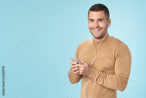 Happy man using mobile phone listening music with earphones posing isolated over blue background