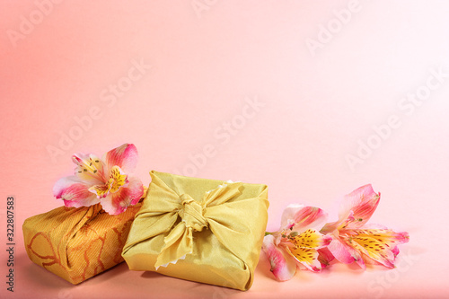 Trendy wrapped in golden textile in Furoshiki technique gift boxes and flowers Alstroemeria on pink. Valentine's day, mother's day, Women's day background. Zero waste holiday. Copy space.