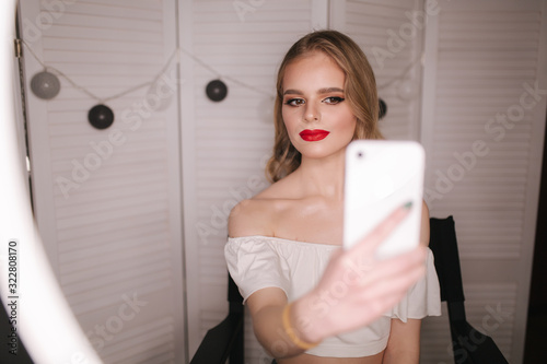 Young attractive model make a selfie after makeup in beauty studio. She use a ring lapm for photo. Background of white folding screen. Red lipstick photo