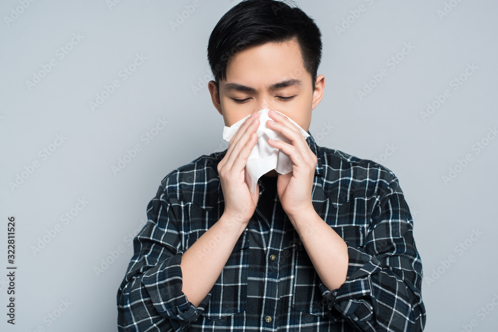 young asian man with closed eyes wiping nose with paper napkin while suffering from runny nose isolated on grey young asian man with closed eyes wiping nose with paper napkin while suffering from runny nose isolated on grey