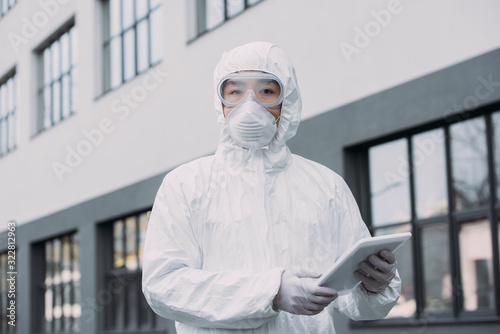 asian epidemiologist in hazmat suit and respirator mask holding digital tablet and looking at camera while standing on street photo