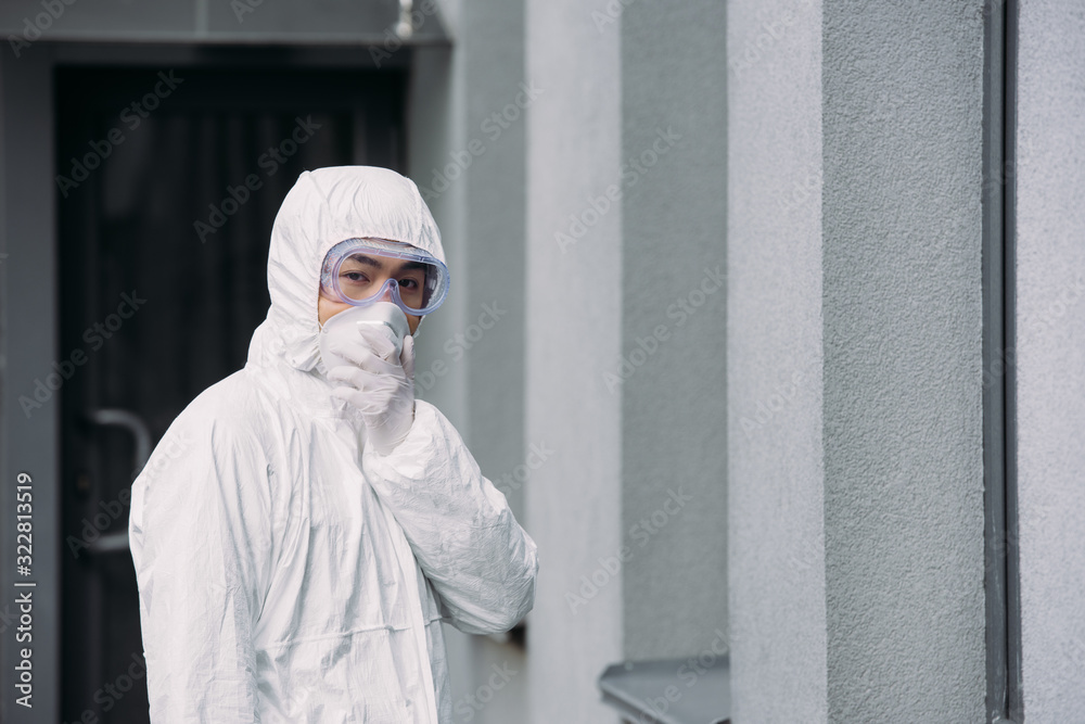 asian epidemiologist in hazmat suit and respirator mask looking at camera while standing outside