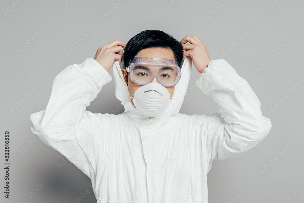 asian epidemiologist in hazmat suit and respirator mask putting hood on while looking at camera on grey background asian epidemiologist in hazmat suit and respirator mask putting hood on while looking at camera on grey background