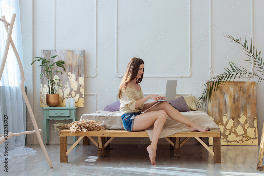 Beautiful lifestyle natural looking picture of woman with long hair sitting by the wooden bed and working on her laptop surrounded by boho light interior Beautiful lifestyle natural looking picture of woman with long hair sitting by the wooden bed and working on her laptop surrounded by boho light interior