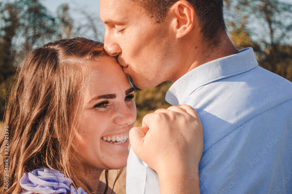 Close up of young couple in love hugging and smiling in hay field. Man hugs woman closely. Man kissing woman in head. Outdoor portrait of beautiful romantic couple in light clothes posing in summer Close up of young couple in love hugging and smiling in hay field. Man hugs woman closely. Man kissing woman in head. Outdoor portrait of beautiful romantic couple in light clothes posing in summer