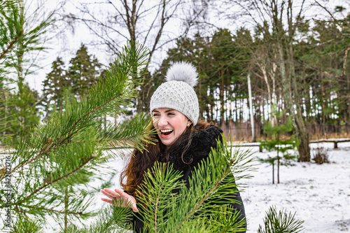 A girl in a light hat and a dark jacket stands among small pines and laughs.