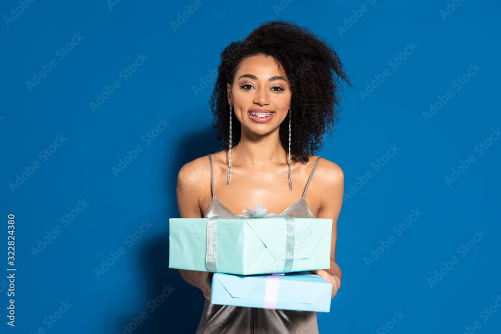 Fototapeta premium smiling beautiful african american woman in silver dress holding gifts on blue background
