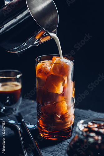Ice coffee from ice cubes in a tall glass  metal pitcher with milk  dark mood  metal straws for cocktails  a jar of coffee beans  black background