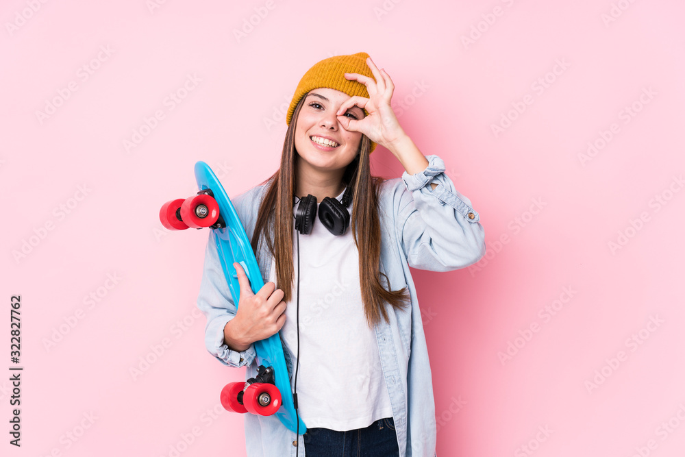 Young skater woman holding a skate excited keeping ok gesture on eye.